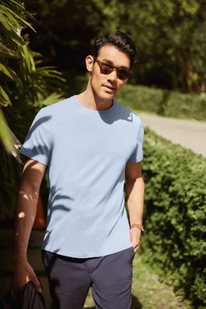 Man wearing a light blue t-shirt and sunglasses walking along an outdoor path lined with lush green foliage.