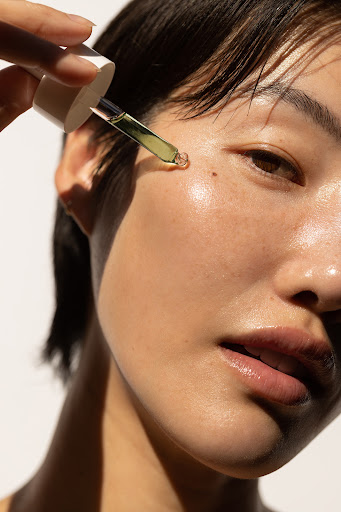 Close-up of a woman applying serum to her face with a dropper. Her skin appears smooth, and the background is a soft white.