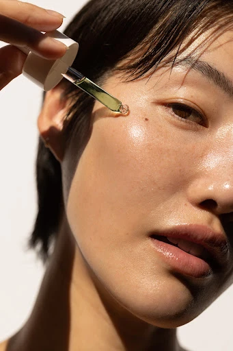 Close-up of a woman applying serum to her face with a dropper. Her skin appears smooth, and the background is a soft white.