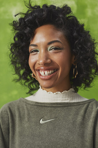 Smiling woman with curly dark hair, wearing a green sweatshirt with a white logo, set against a textured green background.