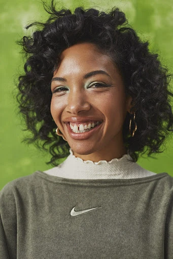 Smiling woman with curly dark hair, wearing a green sweatshirt with a white logo, set against a textured green background.