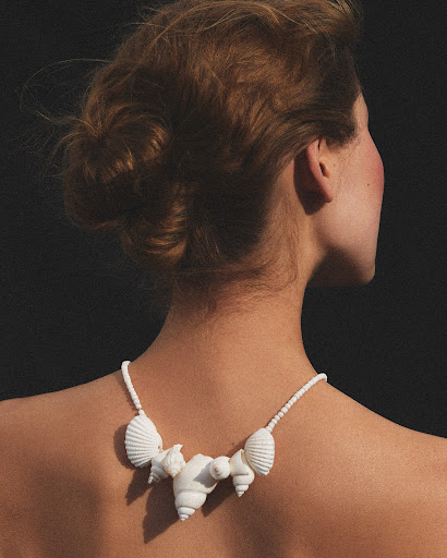 Woman with auburn hair in updo poses from behind wearing a seashell necklace against a dark background in a studio setting.
