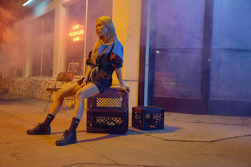 Blonde woman in athletic jacket and shorts sits on black milk crates outside a building with red neon sign at dusk.