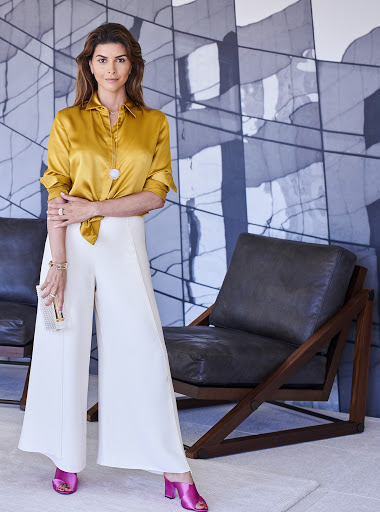 Indoor portrait of a brunette woman in a gold silk blouse and white wide-legged pants standing in front of modern artwork.