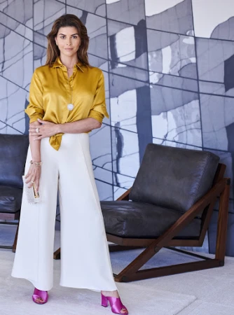 Indoor portrait of a brunette woman in a gold silk blouse and white wide-legged pants standing in front of modern artwork.