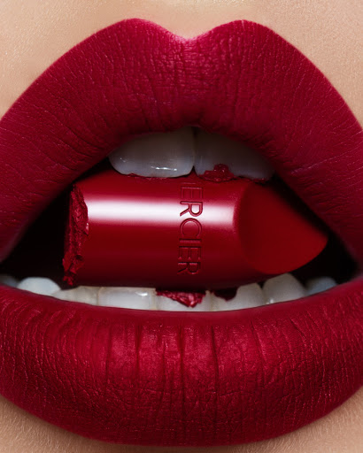 Close-up of bright red matte lipstick applied to lips holding a broken red lipstick tube between white teeth.