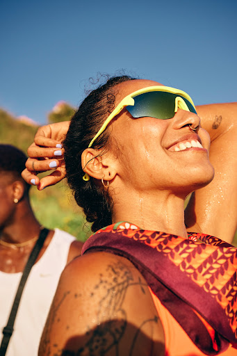 Woman smiles in the sunlight wearing sporty neon sunglasses and a patterned scarf, showcasing fashion and outdoor lifestyle. 