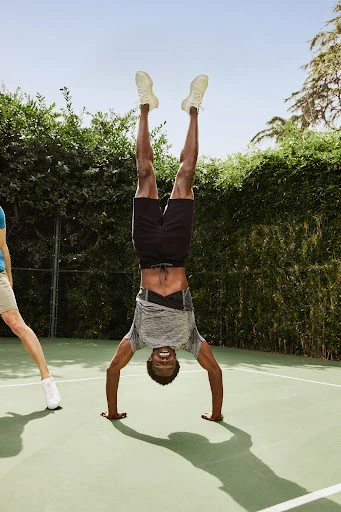 Man in athletic wear performs a handstand on a tennis court with green bushes, blue sky, and another person visible in the background.