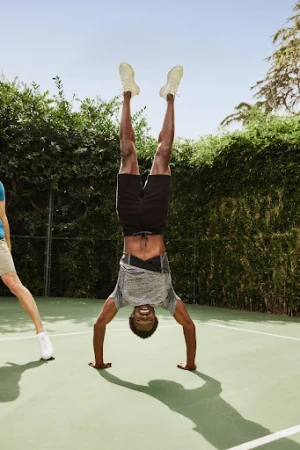 Man in athletic wear performs a handstand on a tennis court with green bushes, blue sky, and another person visible in the background.