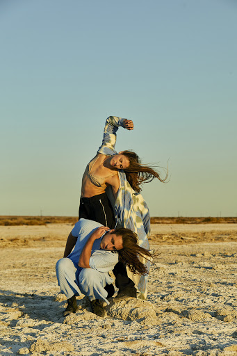 Two adult models pose in a barren landscape with a clear blue sky. One model sits while the other leans back over the first.