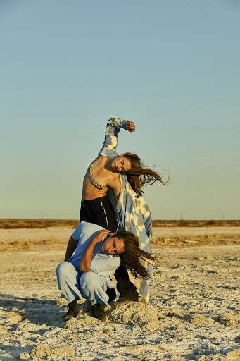 Two adult models pose in a barren landscape with a clear blue sky. One model sits while the other leans back over the first.