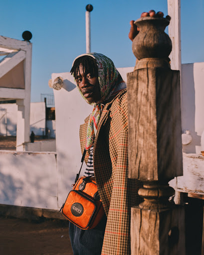 A stylish adult male posing outdoors with a Gucci bag, wearing a patterned headscarf and blazer in front of a white building and wooden post.