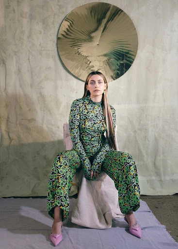 A woman with long hair sits on a chair in a patterned outfit, wearing pink heels, photographed in a studio with a textured backdrop.