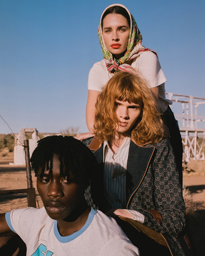 Three fashion models strike a pose in an outdoor setting featuring desert vegetation and a clear blue sky. The group showcases a diverse style.