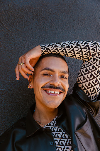 Close-up portrait of a smiling person with a mustache, wearing a patterned top and a black jacket, with a textured gray wall background.