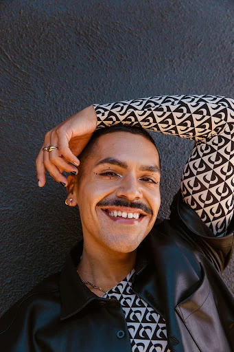 Close-up portrait of a smiling person with a mustache, wearing a patterned top and a black jacket, with a textured gray wall background.