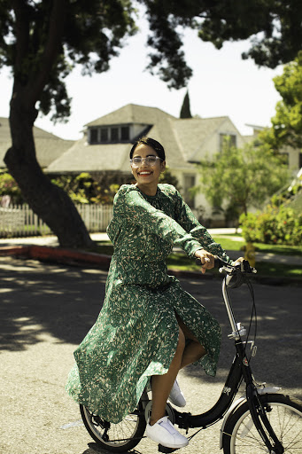 A smiling woman in a green patterned dress is riding a bike on a sunny street, with houses and trees visible in the background.