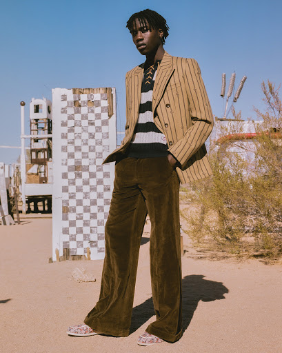 A stylish young Black man poses outdoors in a tan striped blazer, knit shirt, and brown velvet pants against a bright blue sky backdrop.