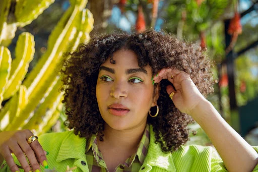 Portrait of an adult woman with curly hair, wearing a lime green jacket, posing outdoors with a cactus and blurred foliage background.