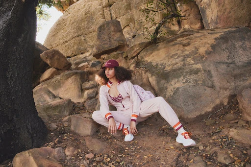 Woman poses in pink sweatsuit on rocky terrain in nature. She wears a cap and athletic shoes in front of large boulders and foliage.