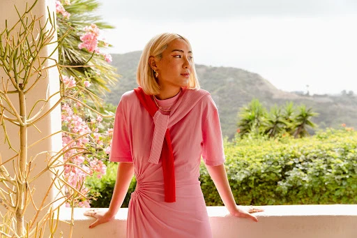 A blonde woman in a pink dress poses on a balcony with foliage and a mountain view, wearing a red scarf and gold jewelry in natural light.