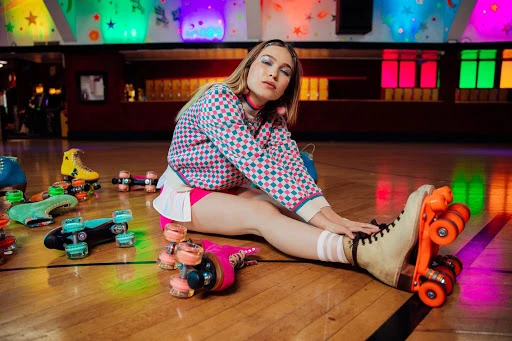 Woman poses with retro roller skates on a wooden floor in a roller rink with colorful lighting and vintage decor, wearing a patterned top.