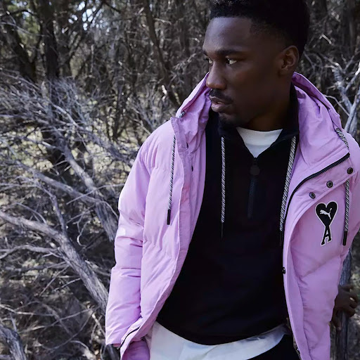 Man in a light purple Puma puffer jacket with black zip-up sweater, captured outdoors against a backdrop of barren winter trees.