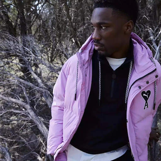 Man in a light purple Puma puffer jacket with black zip-up sweater, captured outdoors against a backdrop of barren winter trees.