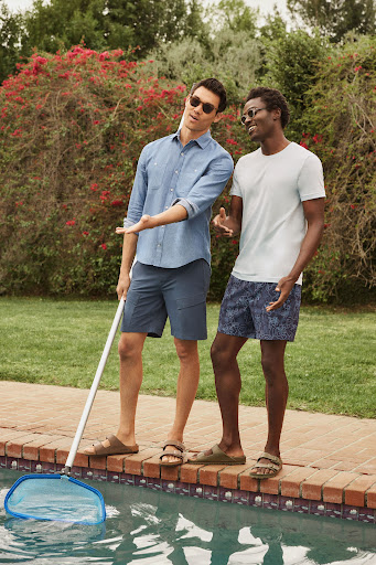 Two men stand poolside in casual summer attire, one holding a pool net, with a green lawn and red-flowered bushes in the background.