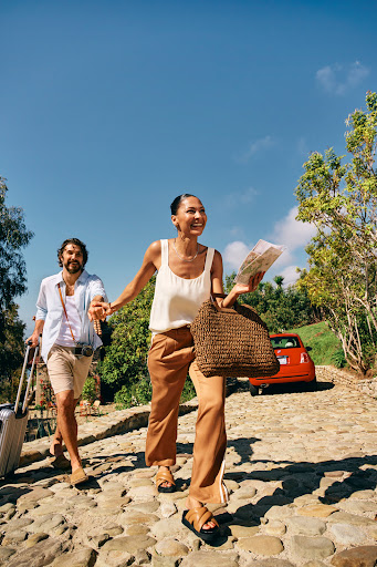 Couple walks hand-in-hand on stone path with luggage, map, and purse. Orange car is parked near trees under a sunny blue sky.