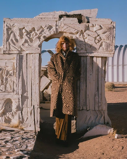 A person in a patterned coat stands in a stone archway ruin in the desert under a blue sky. Fashion, outdoors, daytime.