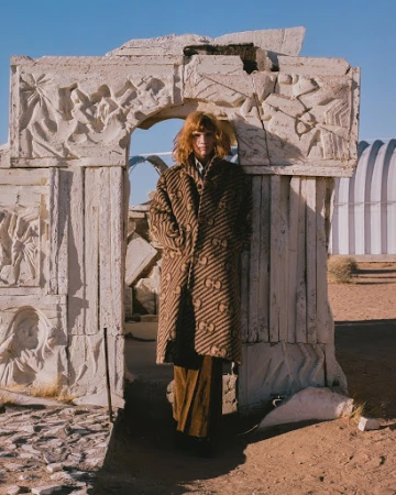 A person in a patterned coat stands in a stone archway ruin in the desert under a blue sky. Fashion, outdoors, daytime.