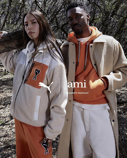 Two models showcase Puma x Ami apparel in an outdoor setting with trees. The woman wears a zip-up jacket and sweatpants; the man is wearing a coat.