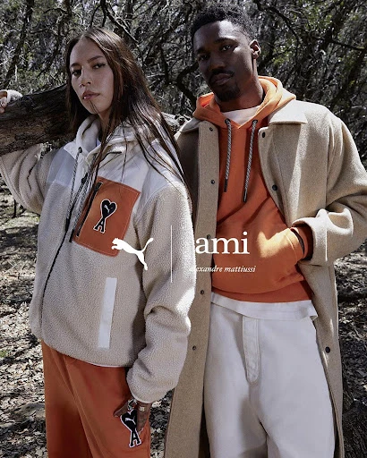 Two models showcase Puma x Ami apparel in an outdoor setting with trees. The woman wears a zip-up jacket and sweatpants; the man is wearing a coat.