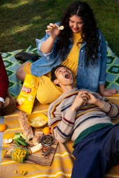 Woman holding a crisp while resting her hand above a man lying on a picnic blanket with snacks and a bag of chips