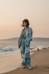Woman in blue patterned suit standing barefoot on a sandy beach at sunset with calm ocean waves and seagulls in the background.