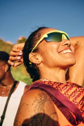Smiling woman wearing yellow athletic sunglasses and patterned scarf with makeup by Omayma Ramzy under clear blue sky.