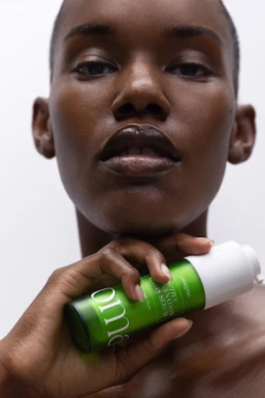 Close-up portrait of a woman holding a green skincare bottle near her chin against a white background, with makeup by Omayma Ramzy.