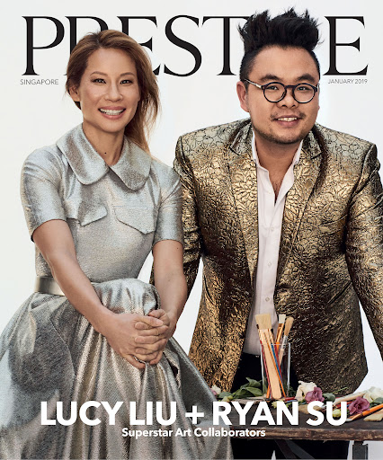Magazine cover featuring Lucy Liu and Ryan Su, artists, in a close-up shot. Liu wears a metallic dress and Su wears a gold brocade blazer.