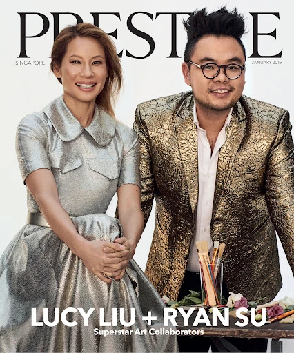 Magazine cover featuring Lucy Liu and Ryan Su, artists, in a close-up shot. Liu wears a metallic dress and Su wears a gold brocade blazer.