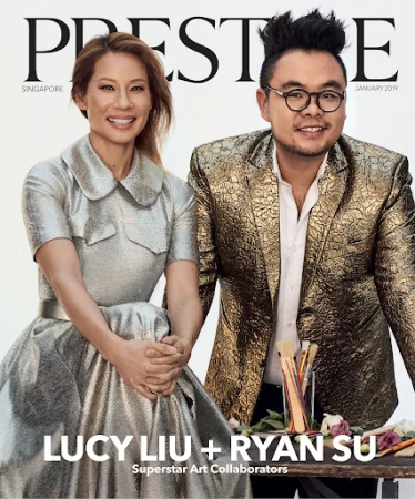 Magazine cover featuring Lucy Liu and Ryan Su, artists, in a close-up shot. Liu wears a metallic dress and Su wears a gold brocade blazer.