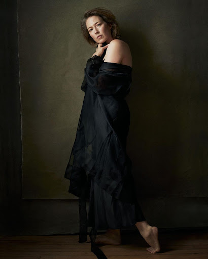 Adult woman poses barefoot wearing a sheer black gown in a studio against a neutral backdrop with dark wood flooring.