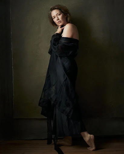 Adult woman poses barefoot wearing a sheer black gown in a studio against a neutral backdrop with dark wood flooring.