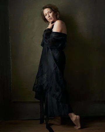 Adult woman poses barefoot wearing a sheer black gown in a studio against a neutral backdrop with dark wood flooring.