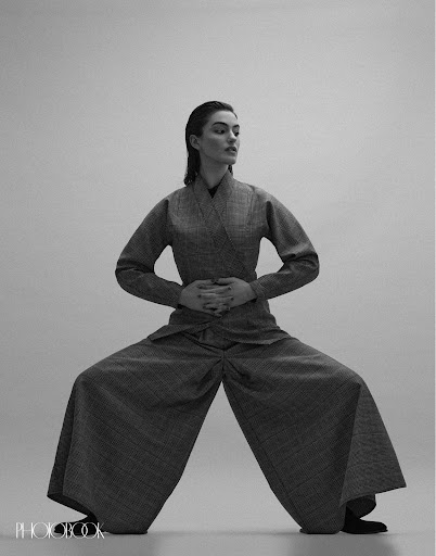 Monochrome studio shot of a young woman posing in a wide-legged pantsuit with hands clasped at the waist, in a neutral grey setting.
