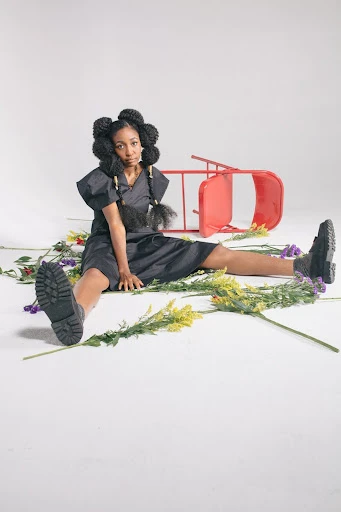 Young African-descent woman in a black dress and boots sits on the floor surrounded by flowers with a red chair in the background.