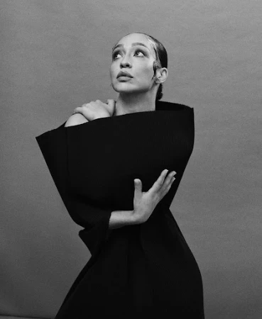Monochrome portrait of a woman wearing a structured black sculptural garment with styled wet-look hair against a grey backdrop.
