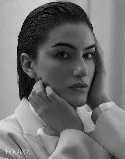 Black and white portrait of a woman with slicked-back hair, wearing a light-colored blazer, and posing with hands near her face.