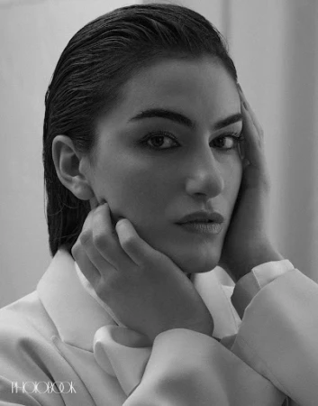 Black and white portrait of a woman with slicked-back hair, wearing a light-colored blazer, and posing with hands near her face.