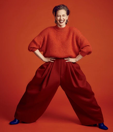A smiling woman stands in front of an orange background wearing a red sweater and wide-leg pants, accessorized with large hoop earrings.